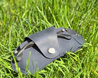 May include: A dark blue leather glasses case with a silver snap closure, holding a pair of eyeglasses. The case is resting in a bed of green grass, with the glasses partially visible.
