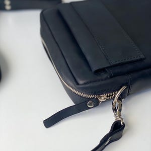 May include: Black leather crossbody bag with a silver zipper and hardware. The bag has a rectangular shape with a flap and a long, adjustable strap. The bag is made of smooth, high-quality leather.