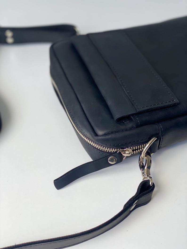 May include: Black leather crossbody bag with a silver zipper and hardware. The bag has a rectangular shape with a flap and a long, adjustable strap. The bag is made of smooth, high-quality leather.