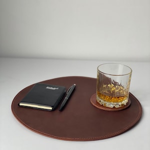 May include: A brown leather desk mat with a black notebook, pen, and a rocks glass with amber liquid on a coaster. The notebook has the word "Notebooks" printed on it.