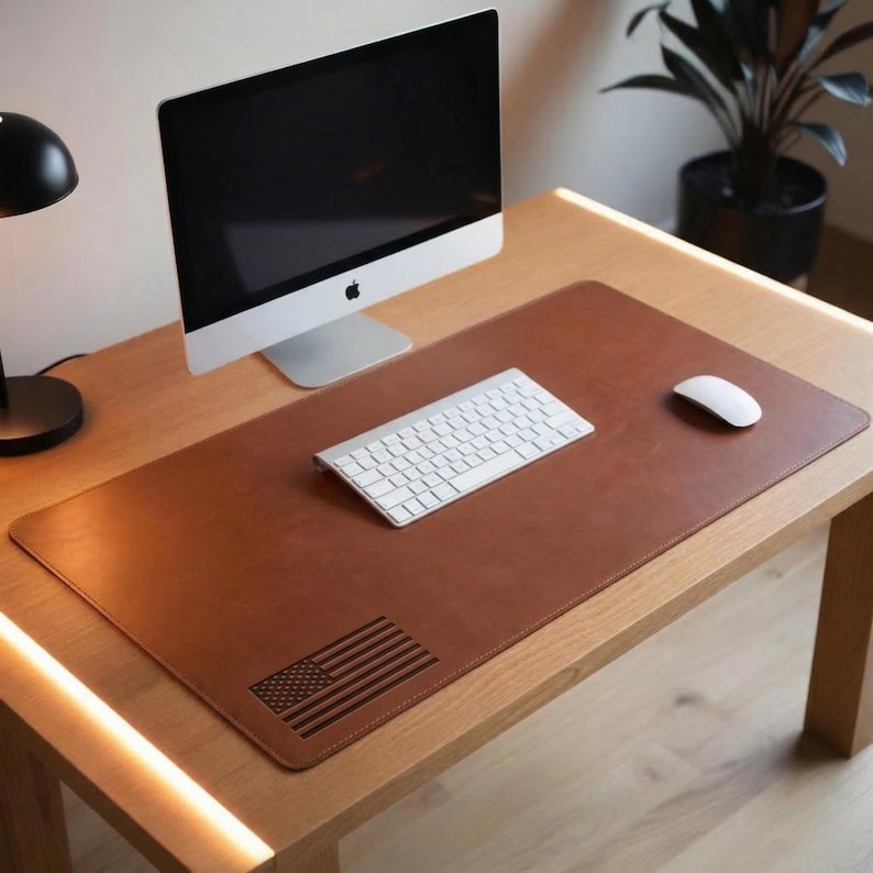 May include: A brown leather desk mat featuring an engraved American flag. A white keyboard and mouse are on the mat, with a silver computer monitor behind. The desk is made of light wood. The mat is approximately 80 cm long.