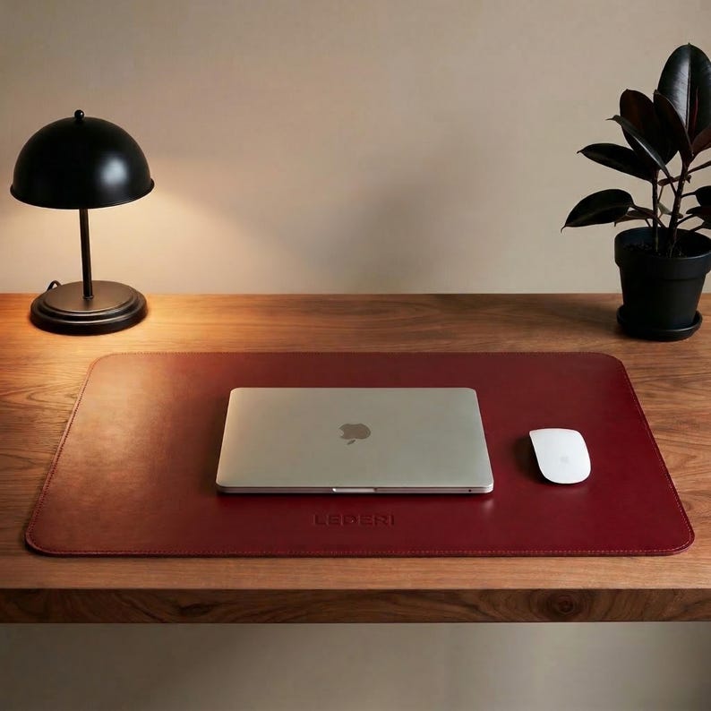 May include: A burgundy leather desk mat with a laptop, mouse, and a black desk lamp. The mat has the word "LEDERI" embossed on it. A potted plant sits on the wooden desk.