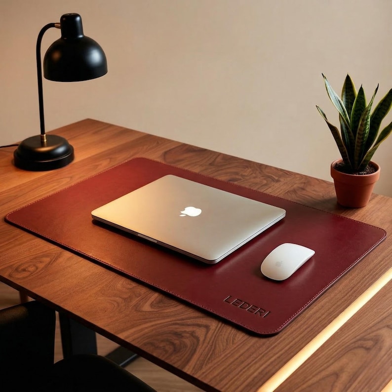 May include: A burgundy leather desk mat with a laptop, mouse, and a small potted plant on a wooden desk. A black desk lamp is on the left. The mat has the word "LEDERI" embossed on the bottom right.