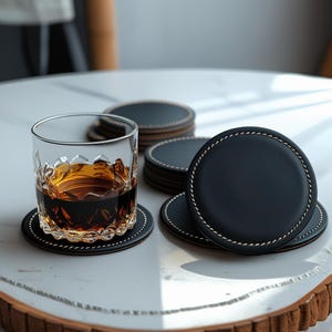 Könnte beinhalten: Ein Glas mit bernsteinfarbener Flüssigkeit steht auf einem schwarzen Lederuntersetzer mit weißen Nähten. Mehrere ähnliche Untersetzer sind in der Nähe gestapelt. Die Untersetzer sind rund und haben einen braunen Rand.
