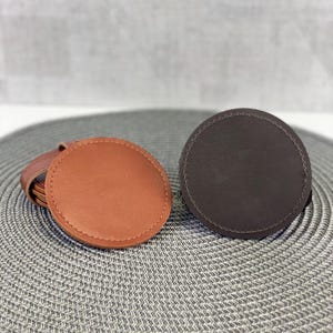 Puede incluir: Dos posavasos redondos de cuero, uno marrón y otro marrón oscuro, sobre una superficie gris texturizada.