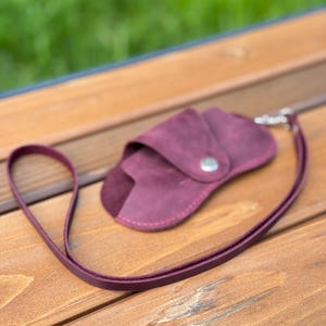 May include: A burgundy leather glasses case with a matching lanyard. The case has a snap closure and a silver-coloured metal clasp. The case is made of a soft, textured leather and is on a wooden surface.