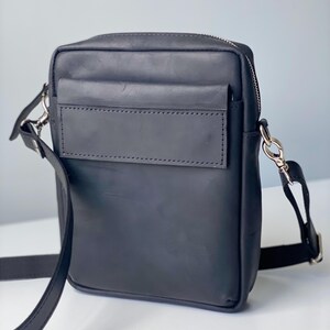 May include: A black leather crossbody bag with a rectangular shape. It features a front pocket, a zipper closure, and a long adjustable strap. The bag has silver-colored hardware.