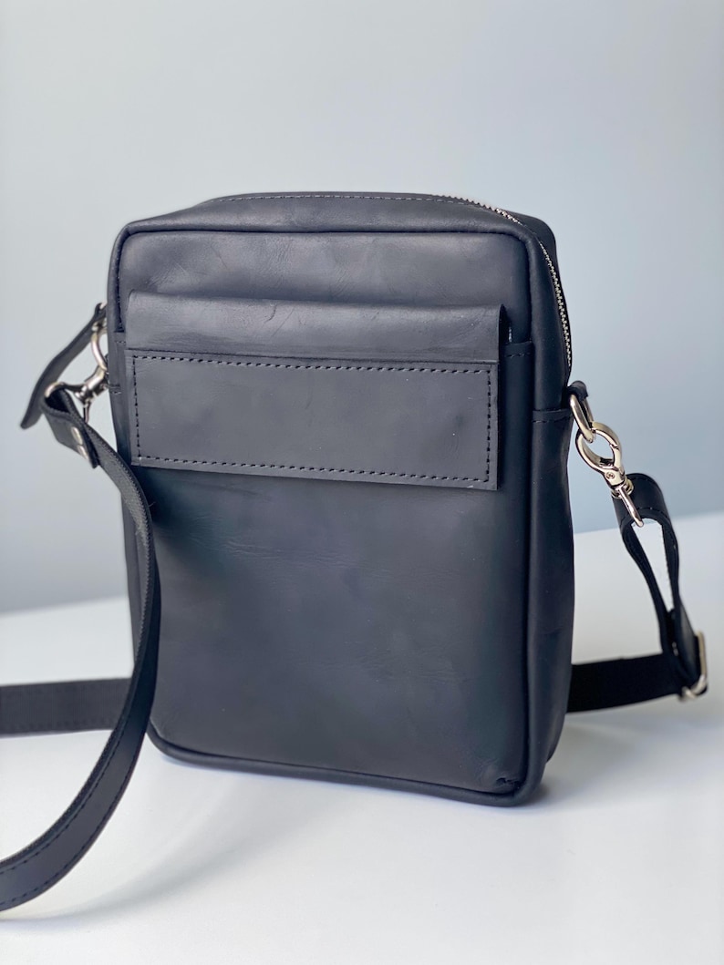May include: A black leather crossbody bag with a rectangular shape. It features a front pocket, a zipper closure, and a long adjustable strap. The bag has silver-colored hardware.