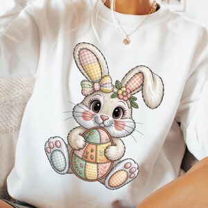 May include: White sweatshirt featuring a cartoon Easter bunny holding a colorful egg. The bunny has a bow, flower crown, and pink cheeks. The egg is decorated with pastel patchwork patterns, perfect for Easter.