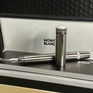 Peut inclure: Un stylo plume Montblanc argent&eacute; avec un dessus blanc, pr&eacute;sent&eacute; dans un coffret noir ouvert. Le stylo repose sur un carnet noir avec le logo Montblanc. La plume du stylo est visible, et le coffret a un couvercle textur&eacute;.