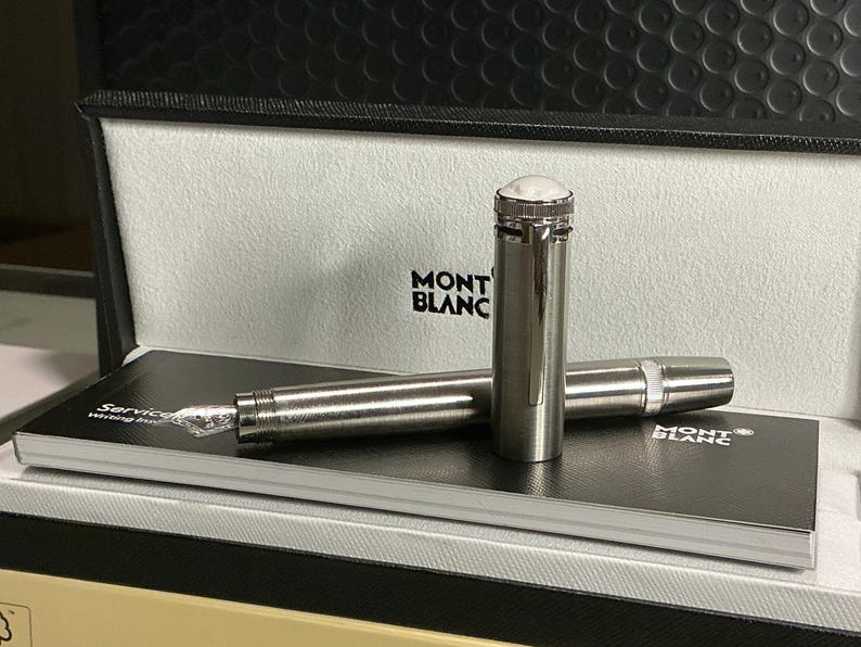 Peut inclure: Un stylo plume Montblanc argent&eacute; avec un dessus blanc, pr&eacute;sent&eacute; dans un coffret noir ouvert. Le stylo repose sur un carnet noir avec le logo Montblanc. La plume du stylo est visible, et le coffret a un couvercle textur&eacute;.