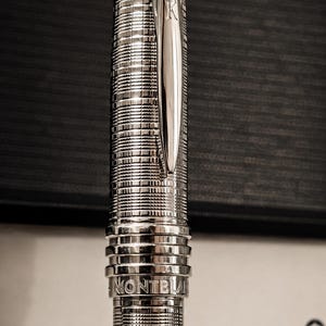May include: A silver-toned fountain pen with a textured, grid-like pattern on the barrel and cap. The pen features a polished clip, a band engraved with the word MONTBLANC, and a white emblem on the top. The brand name MONTBLANC is visible nearby.