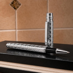 May include: A silver rollerball pen with a repeating oval pattern engraved on the barrel and cap. The cap stands upright next to the pen body, which features a black grip section and a metallic tip. The clip on the cap is engraved with the word Cartier.