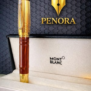May include: A gold-coloured pen with a brown wooden barrel, displayed in a Montblanc box. The pen has a chain and is set against a textured black background with the Penora logo.