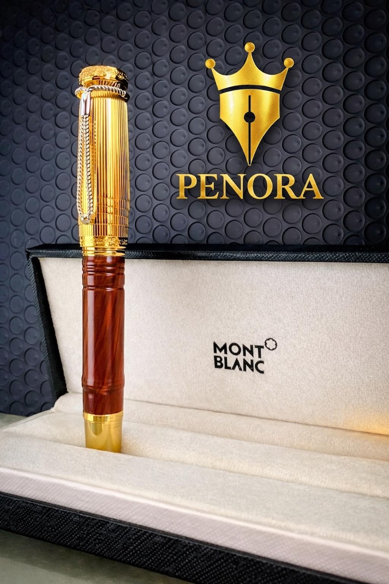 May include: A gold-coloured pen with a brown wooden barrel, displayed in a Montblanc box. The pen has a chain and is set against a textured black background with the Penora logo.