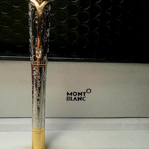 May include: A Montblanc fountain pen with a gold-colored crown and base, and a silver-colored barrel with an ornate floral design. The pen is displayed in a white box with the Montblanc logo.