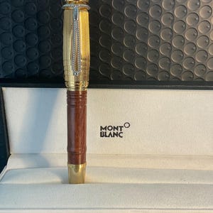 May include: A gold Montblanc fountain pen with a brown wooden barrel, displayed in a white box. The pen has detailed engraving on the cap and a silver chain. The Montblanc logo is printed on the box.