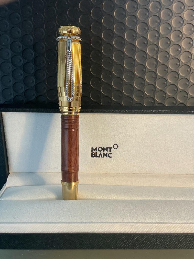 May include: A gold Montblanc fountain pen with a brown wooden barrel, displayed in a white box. The pen has detailed engraving on the cap and a silver chain. The Montblanc logo is printed on the box.