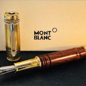 May include: A luxury Montblanc fountain pen with a gold-plated barrel and a dark wood grip. The pen's cap is gold with a white emblem. The pen is displayed with its cap off, revealing the nib. The Montblanc logo is visible.