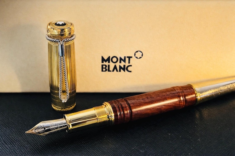 May include: A luxury Montblanc fountain pen with a gold-plated barrel and a dark wood grip. The pen's cap is gold with a white emblem. The pen is displayed with its cap off, revealing the nib. The Montblanc logo is visible.