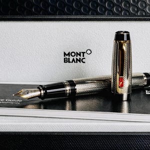 May include: A Montblanc fountain pen with a silver and gold finish, presented in a black and white box. The pen features a detailed engraved pattern and a red accent. A service guide is visible.