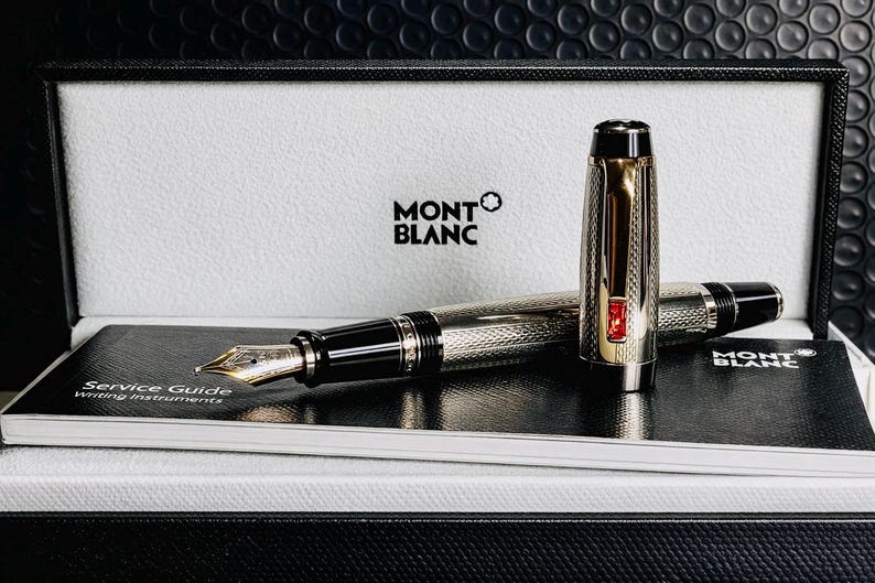May include: A Montblanc fountain pen with a silver and gold finish, presented in a black and white box. The pen features a detailed engraved pattern and a red accent. A service guide is visible.