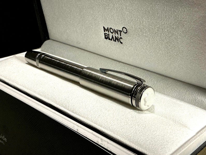Peut inclure: Un stylo Montblanc argent&eacute; est pr&eacute;sent&eacute; dans un coffret noir avec un int&eacute;rieur blanc. Le stylo a une finition m&eacute;tallique brillante, un clip et un embl&egrave;me blanc. Le logo Montblanc est visible sur le coffret.