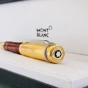 May include: A gold Montblanc pen with a brown leather grip, displayed in a white box. The pen has a fluted gold barrel, silver detailing, and the Montblanc emblem on the end.