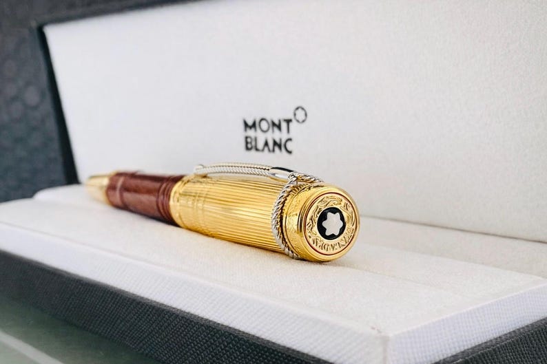 May include: A gold Montblanc pen with a brown leather grip, displayed in a white box. The pen has a fluted gold barrel, silver detailing, and the Montblanc emblem on the end.