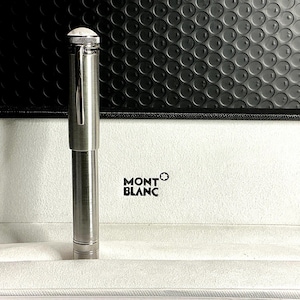 Peut inclure: Un stylo Montblanc argent&eacute; est pr&eacute;sent&eacute; dans son coffret noir et blanc ouvert. Le stylo est cylindrique, avec un clip argent&eacute; et un sommet blanc. Le logo Montblanc est imprim&eacute; sur la bo&icirc;te.