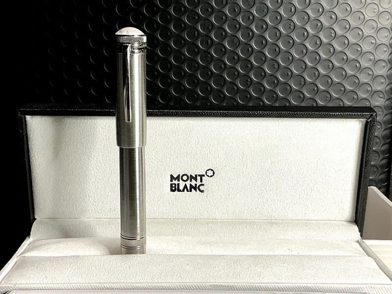 Peut inclure: Un stylo Montblanc argent&eacute; est pr&eacute;sent&eacute; dans son coffret noir et blanc ouvert. Le stylo est cylindrique, avec un clip argent&eacute; et un sommet blanc. Le logo Montblanc est imprim&eacute; sur la bo&icirc;te.