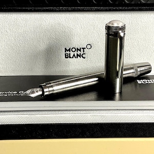 Peut inclure: Un stylo plume Montblanc dans un coffret. Le stylo est argent&eacute; avec un dessus blanc et une plume. La bo&icirc;te porte le logo Montblanc et un int&eacute;rieur noir et blanc. Un guide de service est visible.