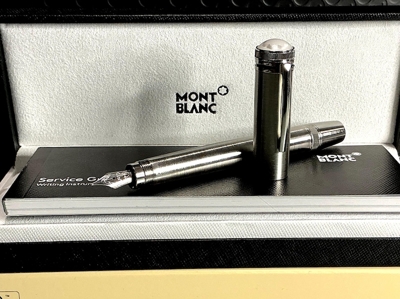 Peut inclure: Un stylo plume Montblanc dans un coffret. Le stylo est argent&eacute; avec un dessus blanc et une plume. La bo&icirc;te porte le logo Montblanc et un int&eacute;rieur noir et blanc. Un guide de service est visible.