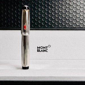 May include: A silver Montblanc pen with a textured design, a red accent, and a black tip, presented in a white box with the Montblanc logo. The pen is standing upright, showcasing its sleek design.