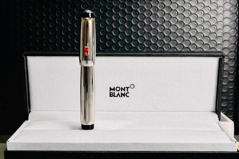 May include: A silver Montblanc pen with a textured design, a red accent, and a black tip, presented in a white box with the Montblanc logo. The pen is standing upright, showcasing its sleek design.