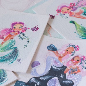 Puede incluir: Cinco ilustraciones de sirenas pintadas a mano con acuarela sobre cartulina blanca. Cada sirena tiene una pose diferente y presenta detalles de purpurina. Las sirenas est&aacute;n rodeadas de flores y otros detalles marinos.