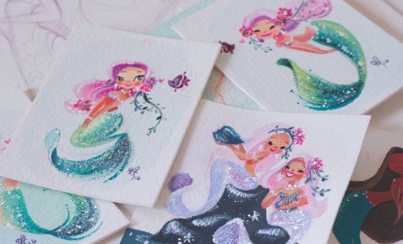 Puede incluir: Cinco ilustraciones de sirenas pintadas a mano con acuarela sobre cartulina blanca. Cada sirena tiene una pose diferente y presenta detalles de purpurina. Las sirenas est&aacute;n rodeadas de flores y otros detalles marinos.