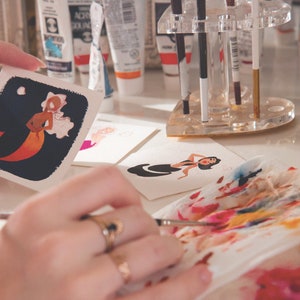 Puede incluir: Una persona est&aacute; pintando una ilustraci&oacute;n de acuarela de una sirena con colores naranja y rosa en una tarjeta blanca. La sirena est&aacute; sobre un fondo azul oscuro con una luna blanca y nubes. La persona est&aacute; usando un pincel y una paleta de colores.