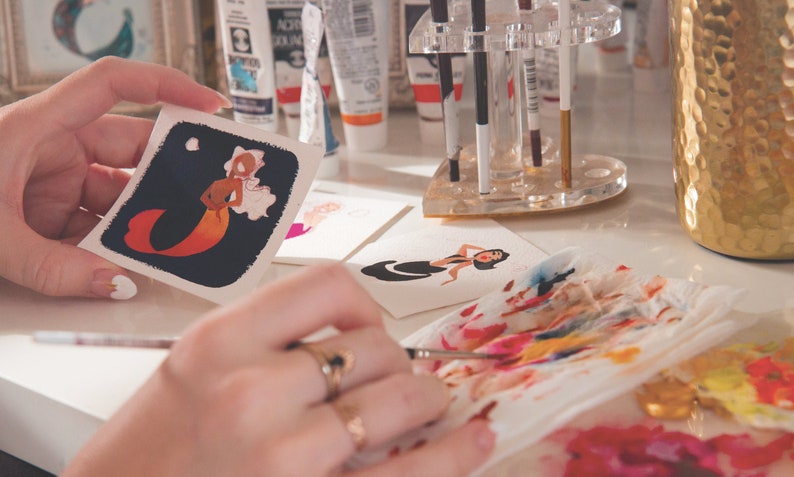 Puede incluir: Una persona est&aacute; pintando una ilustraci&oacute;n de acuarela de una sirena con colores naranja y rosa en una tarjeta blanca. La sirena est&aacute; sobre un fondo azul oscuro con una luna blanca y nubes. La persona est&aacute; usando un pincel y una paleta de colores.