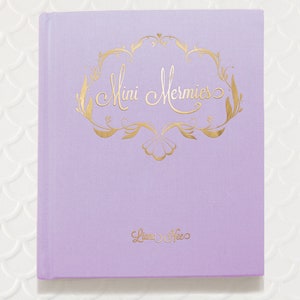 May include: A lavender-colored journal with a gold-colored inscription that reads "Mini Mermies" and "Liana Hee".