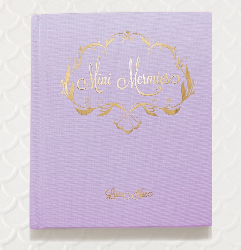 Puede incluir: Un diario de color lavanda con una inscripci&oacute;n dorada que dice "Mini Mermies" y "Liana Hee".