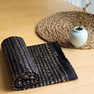 May include: A rolled-up black fabric with gold calligraphy is on a wooden table. A woven placemat and a small white vase with dried plants are in the background. The fabric is partially unrolled, revealing the script.