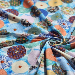 Peut inclure: Gros plan sur un tissu avec un motif répété de parasols colorés et de motifs floraux. Les parasols sont dans des tons d'orange, de bleu, de jaune et de rose, sur un fond bleu clair avec un effet nuageux. Le tissu est plié.
