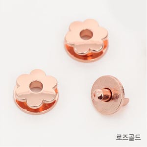 Könnte beinhalten: Drei roségoldene Metall-Druckknöpfe in Blumenform. Zwei der Knöpfe sind flach und haben ein Loch in der Mitte. Der dritte Knopf ist ein Druckverschluss mit einer erhabenen Mitte.