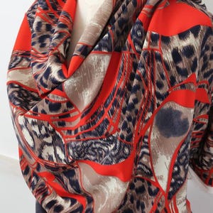 May include: A draped scarf with a bold pattern. The scarf features a vibrant red, tan, and navy blue design with a leopard print and geometric shapes. The fabric is draped over a mannequin, showcasing its flow and texture.