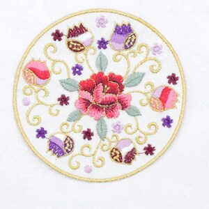 May include: Circular white embroidered patch with a floral design. The patch features a large red and pink flower in the centre, surrounded by smaller flowers in shades of purple and pink, with gold trim.