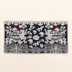 Puede incluir: Parche rectangular bordado con fondo azul oscuro y detalles en blanco y rojo. El diseño presenta dos criaturas míticas estilizadas, nubes y patrones de olas. El parche es probablemente para fines decorativos.