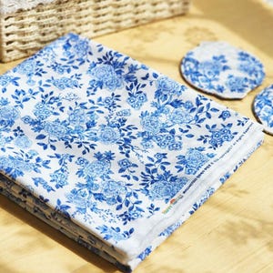 May include: A folded white fabric with a blue floral pattern, alongside two matching coasters. The fabric and coasters feature a repeating design of blue flowers and leaves. The items are arranged on a light-colored wooden surface.