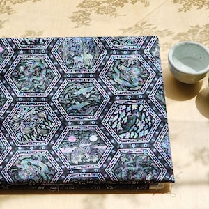 Peut inclure: Un tissu plié à motifs hexagonaux, représentant des créatures mythiques et des paysages en noir, sarcelle et couleurs irisées. Deux petits bols en céramique, un bleu clair et un vert, sont à proximité. Le tissu est posé sur une surface claire.