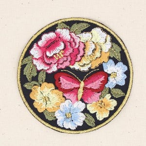 Pode incluir: Remendo bordado circular com um desenho de borboleta e floral. O remendo tem um fundo preto e uma borda dourada. A borboleta é rosa e vermelha, rodeada por flores rosa, amarelas e azuis.